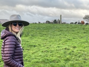 Casey Stevens in Ireland wearing a purple down jacket, sunglasses and a wide brimmed grey hat